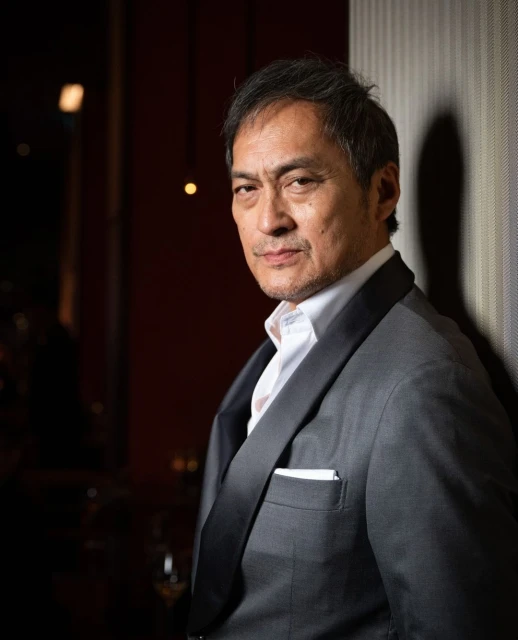 Portrait of a middle-aged adult male wearing a grey suit and a white shirt, looking at the camera in an indoor setting with dark lighting.