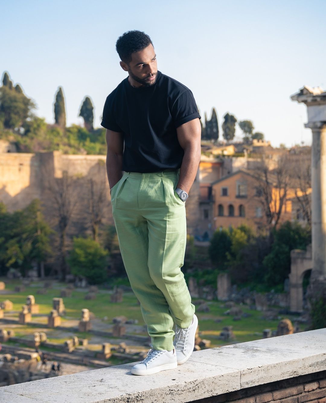 Man in black t-shirt and green pants standing in front of Roman Forum ruins under blue sky, wearing watch, looking away. 