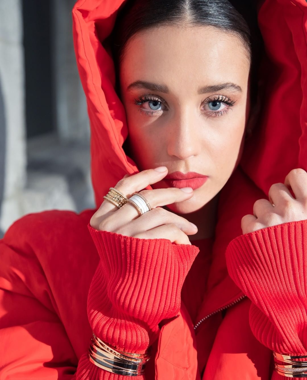 Portrait of a beautiful adult woman with blue eyes wearing a red hooded jacket, rings, and bracelets against a neutral grey background.