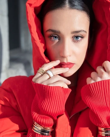 Portrait of a beautiful adult woman with blue eyes wearing a red hooded jacket, rings, and bracelets against a neutral grey background.