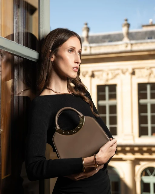 Elegant woman with long hair in black dress holding a taupe handbag by a window, against an architectural backdrop on a sunny day.