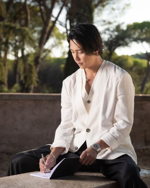 Asian man in a white linen suit sits outdoors, writing in a notebook in a park-like setting with trees in the background.