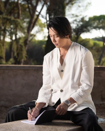 Asian man in a white linen suit sits outdoors, writing in a notebook in a park-like setting with trees in the background.