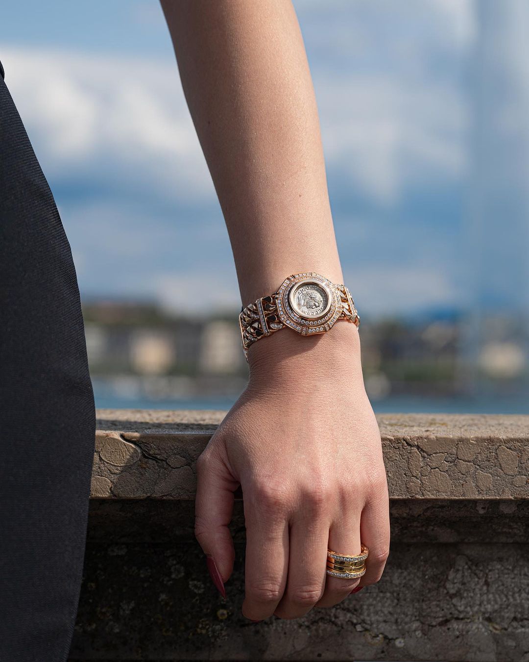 Close-up of a woman's wrist with a gold watch and rings, set against a blurred urban and sky background. Jewellery advertisement.