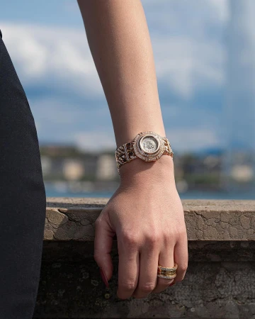 Close-up of a woman's wrist with a gold watch and rings, set against a blurred urban and sky background. Jewellery advertisement.