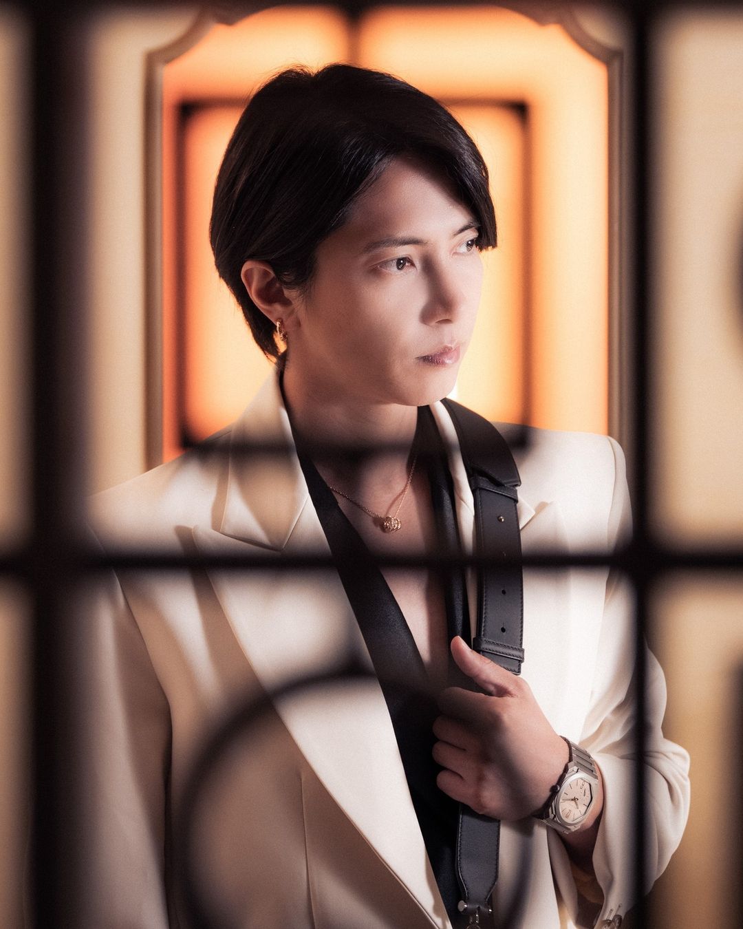 Close-up portrait of a stylish adult male with short dark hair wearing a white suit jacket and watch against a warm backdrop.
