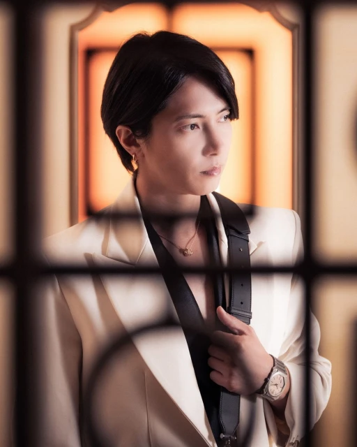 Close-up portrait of a stylish adult male with short dark hair wearing a white suit jacket and watch against a warm backdrop.