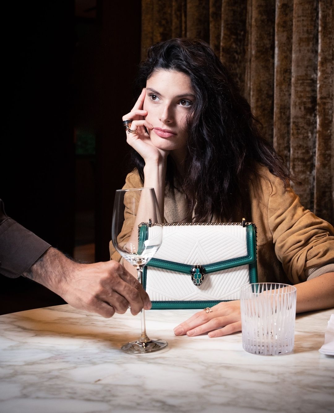 Elegant woman at a table with a drink, featuring a white and green handbag, indoor setting with warm lighting, and marble table.
