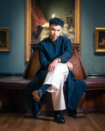 A young man seated on a leather circular bench inside a gallery with framed artwork on dark walls.