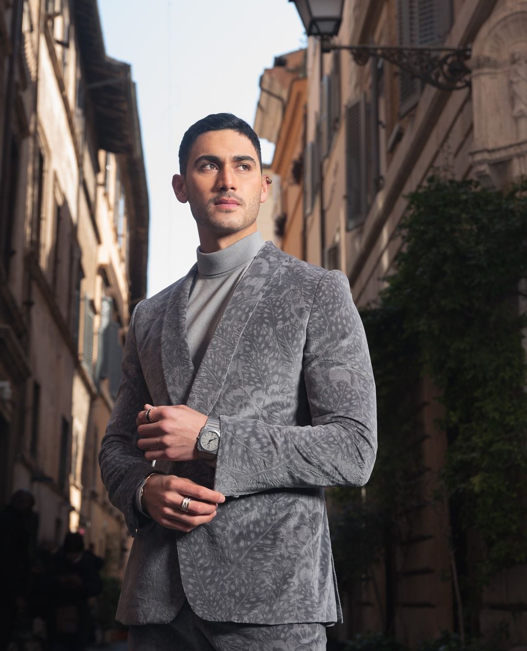 Handsome man in gray patterned suit poses in a European alleyway. He has dark hair, olive skin and wears a gray turtleneck.