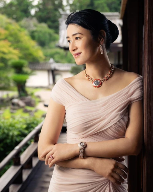 Elegant Asian woman in a light pink dress and jewelry stands on a balcony overlooking a lush, green, Asian garden landscape.