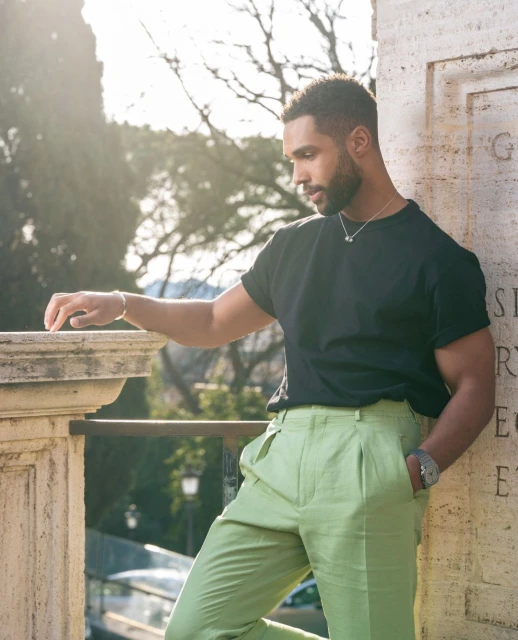 Portrait of a handsome adult male leaning against a stone wall outdoors wearing a black t-shirt and green pants on a sunny day.