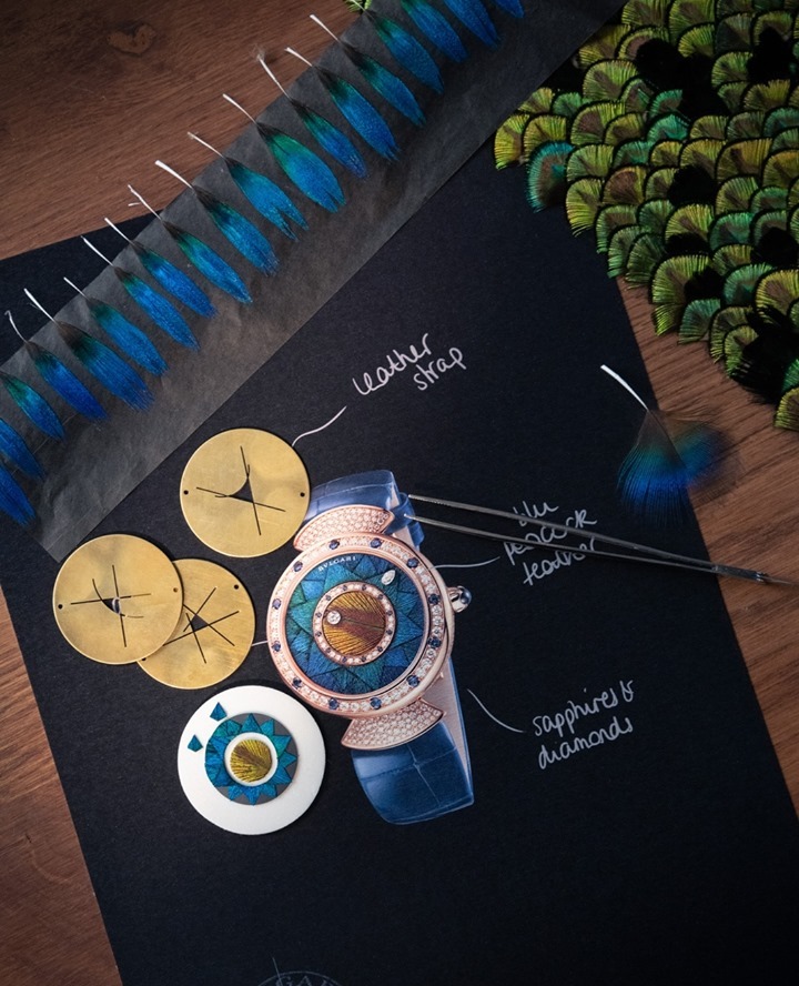 Close-up of a luxury watch design featuring peacock feathers, leather straps, sapphires, and diamonds on a dark surface with gold details.