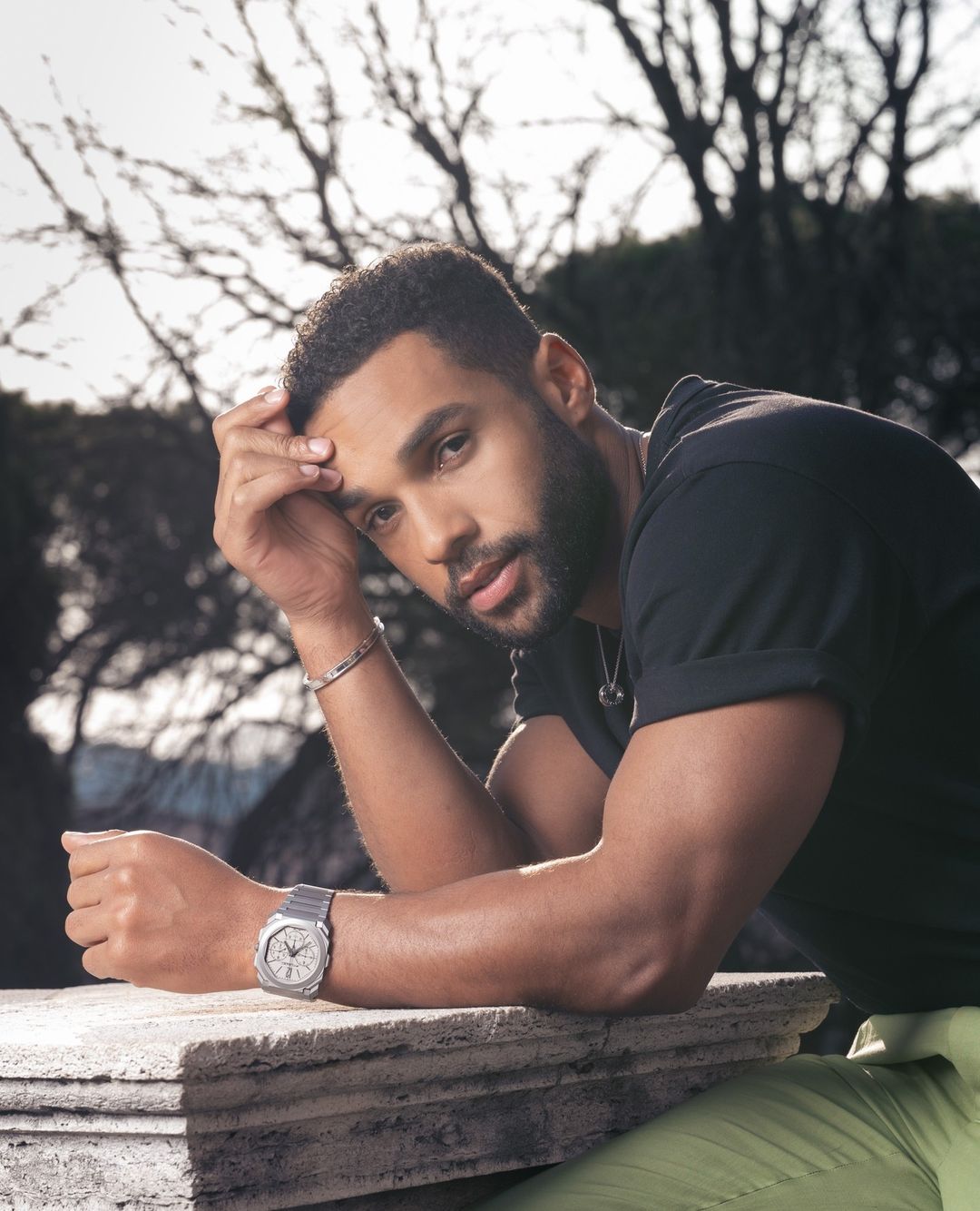 Adult male poses outdoors in natural light wearing a watch and casual black t-shirt. Focus on portrait with a background of trees.