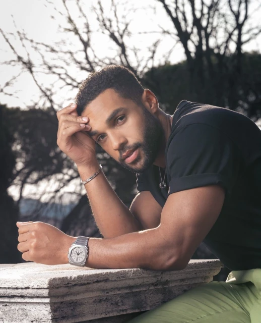 Adult male poses outdoors in natural light wearing a watch and casual black t-shirt. Focus on portrait with a background of trees.