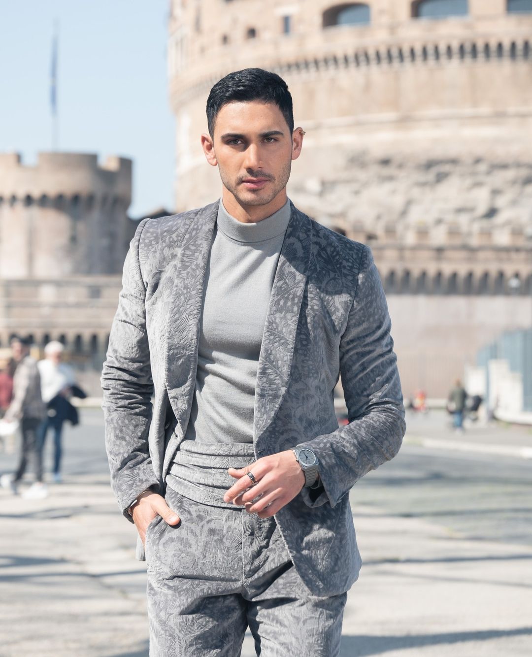 A stylish man in a grey patterned suit and turtleneck stands outdoors with a historic building in the background on a sunny day.
