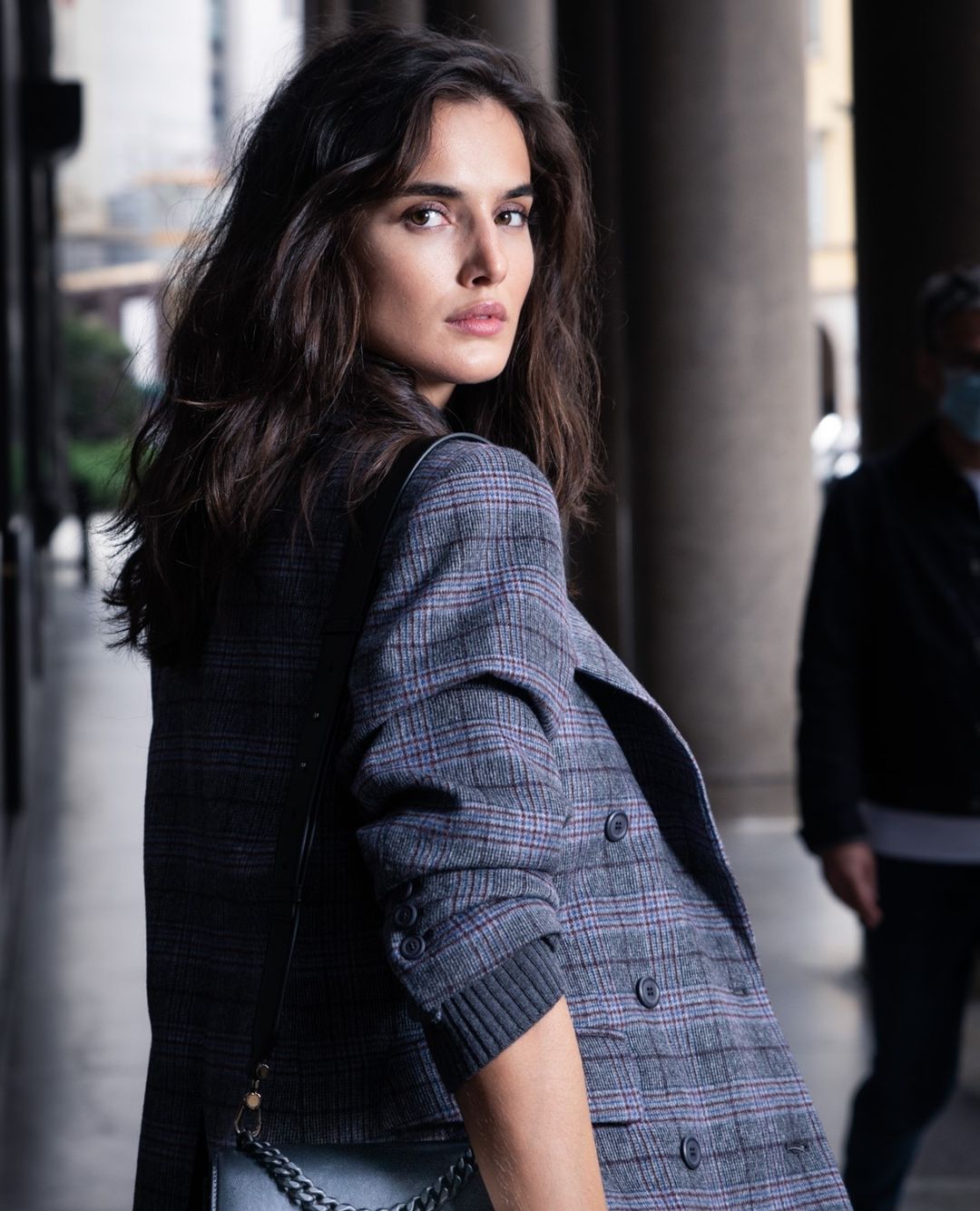Fashionable brunette woman in a grey plaid blazer glances over her shoulder in an urban outdoor setting with neutral-toned columns.