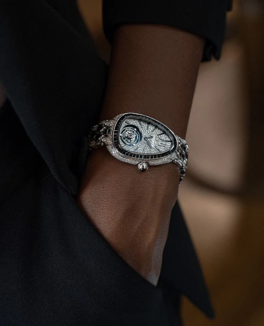 Luxury diamond bracelet watch on an adult's dark skin, with a black blazer, shot indoors with neutral backgrounds and eye-level angle.