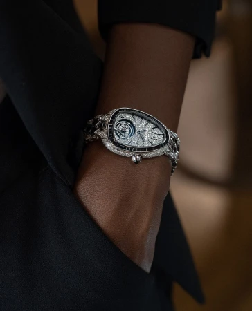 Luxury diamond bracelet watch on an adult's dark skin, with a black blazer, shot indoors with neutral backgrounds and eye-level angle.