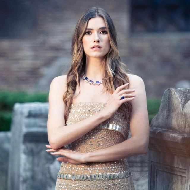 Woman in a golden strapless dress poses outdoors wearing a blue and silver necklace and ring. She has long brown hair.