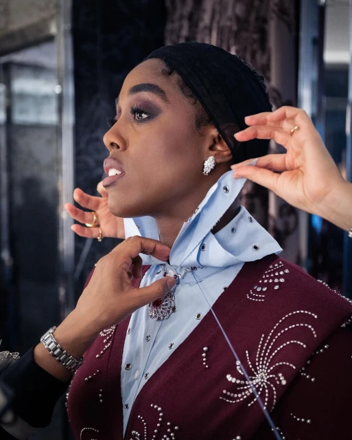 Profile shot of an African-American woman getting ready; stylists adjust her collar and necklace, showcasing fashion and beauty details.