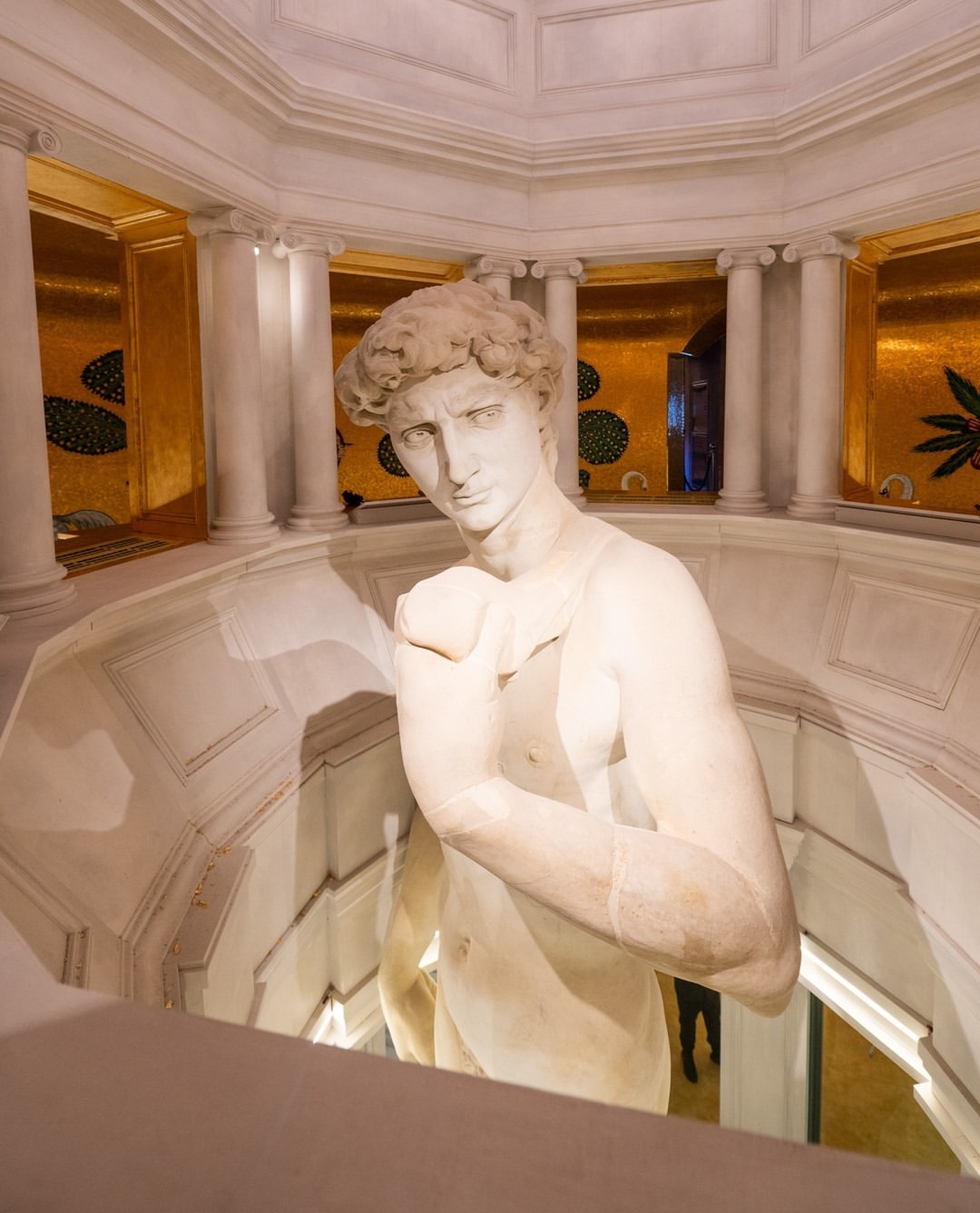 Full-body view of the marble statue of a nude man, posing inside a decorative interior with gold accents and white columns.