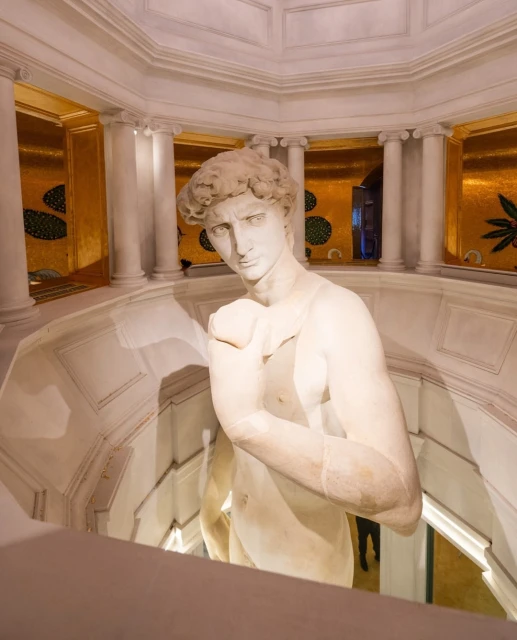 Full-body view of the marble statue of a nude man, posing inside a decorative interior with gold accents and white columns.