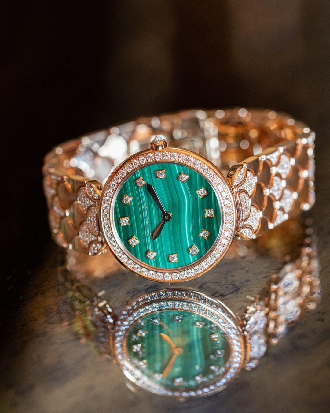 Elegant rose gold wristwatch with malachite face and diamond accents reflected on a dark glossy surface in a close-up studio shot.
