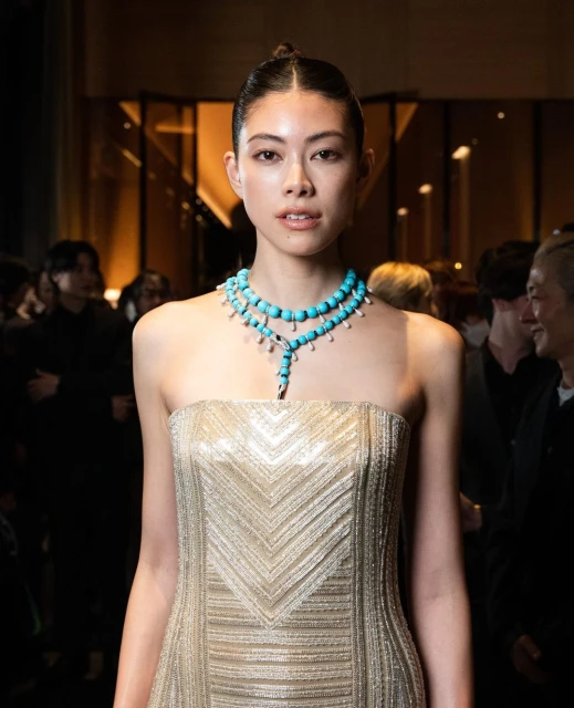 Portrait of a young Asian woman with a high bun, wearing a strapless gold gown and turquoise beaded necklace, in an indoor setting.