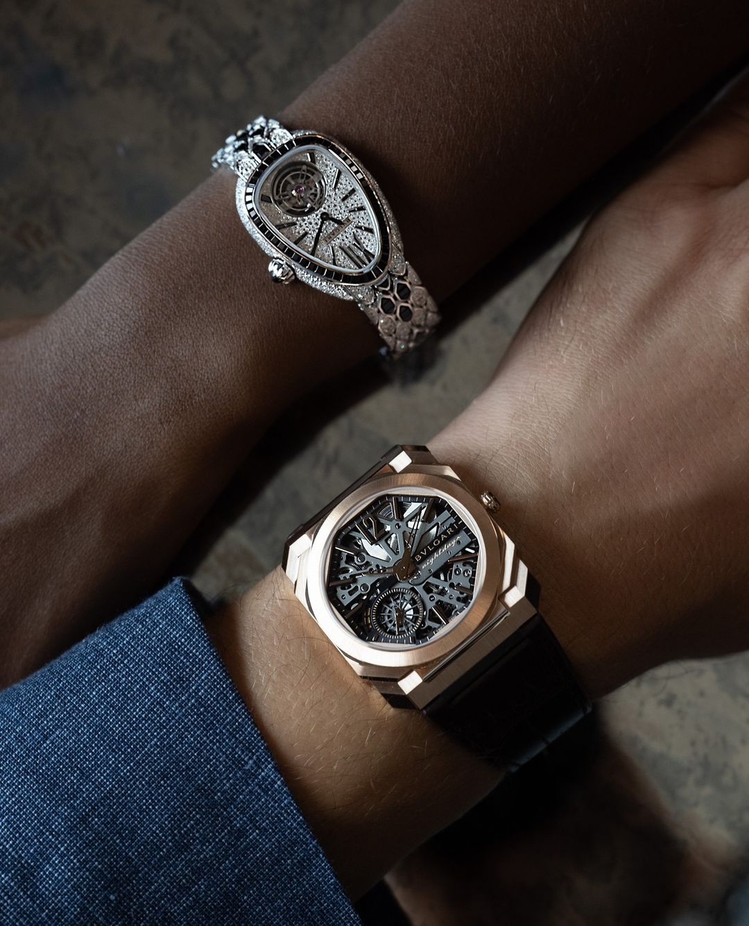 Close-up shot of two people's arms, each adorned with luxury watches. One watch is gold with a black strap, the other is diamond-encrusted.
