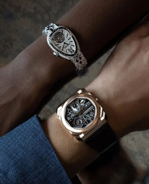 Close-up shot of two people's arms, each adorned with luxury watches. One watch is gold with a black strap, the other is diamond-encrusted.