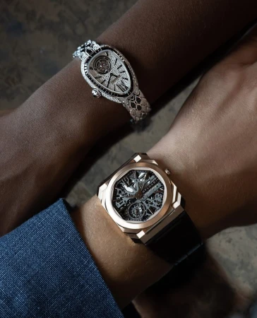Close-up shot of two people's arms, each adorned with luxury watches. One watch is gold with a black strap, the other is diamond-encrusted.