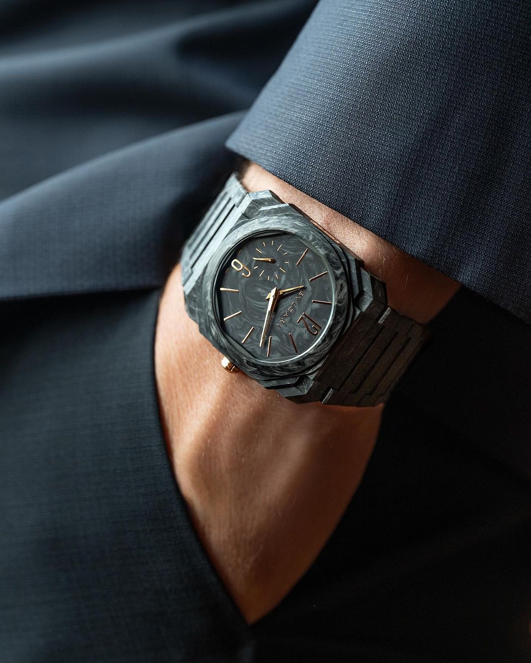 Close-up of an adult hand in a navy suit jacket with a modern black and gray patterned wristwatch with bronze hands and accents.