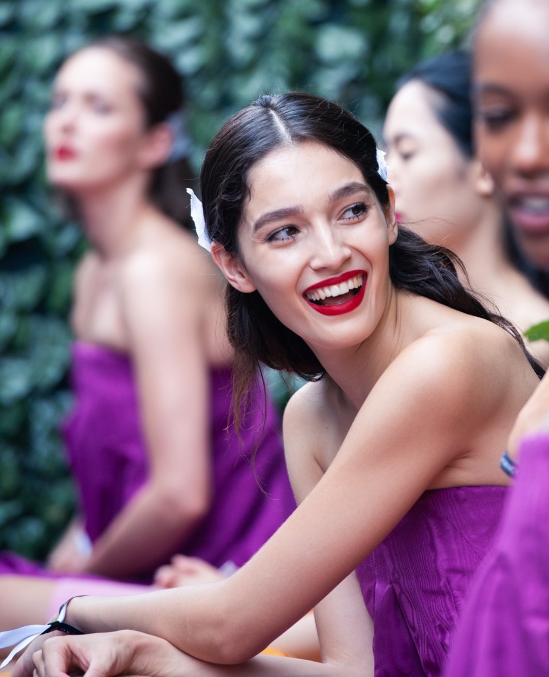 A group of women with red lipstick and flowers in their hair sit together, wrapped in purple towels, outside with a leafy background.