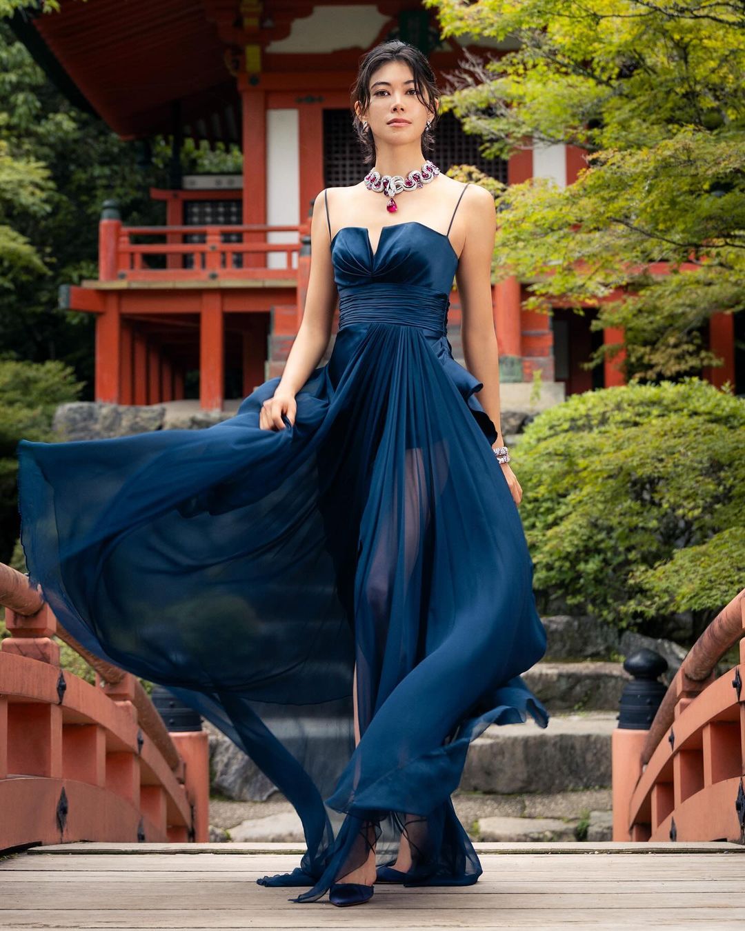 Elegant woman in a blue gown standing on a wooden bridge with a Japanese building and greenery in the background, jewelry details.