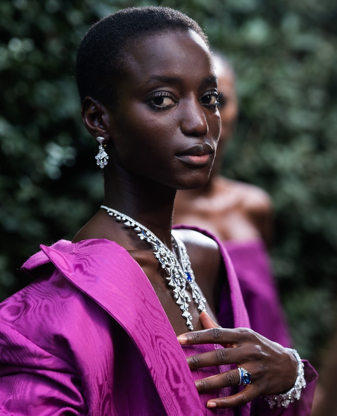 Close-up portrait of a dark-skinned woman wearing formal jewelry and a purple jacket, with a blurry second woman in the background.