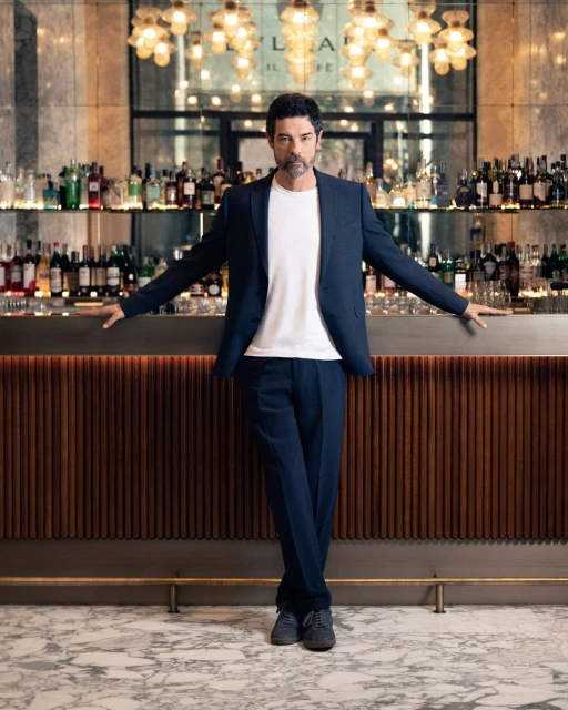 Man in suit leans against bar with liquor bottles at Bulgari Il Ristorante Luca Fantin; portrait format, eye-level angle, natural lighting.