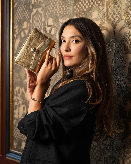 Brunette woman in a black jacket posing indoors with a gold clutch, against a patterned wall, creating an elegant fashion portrait.
