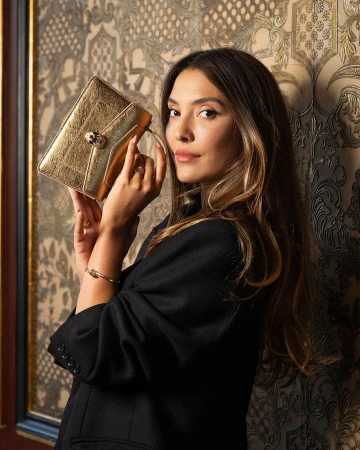 Brunette woman in a black jacket posing indoors with a gold clutch, against a patterned wall, creating an elegant fashion portrait.