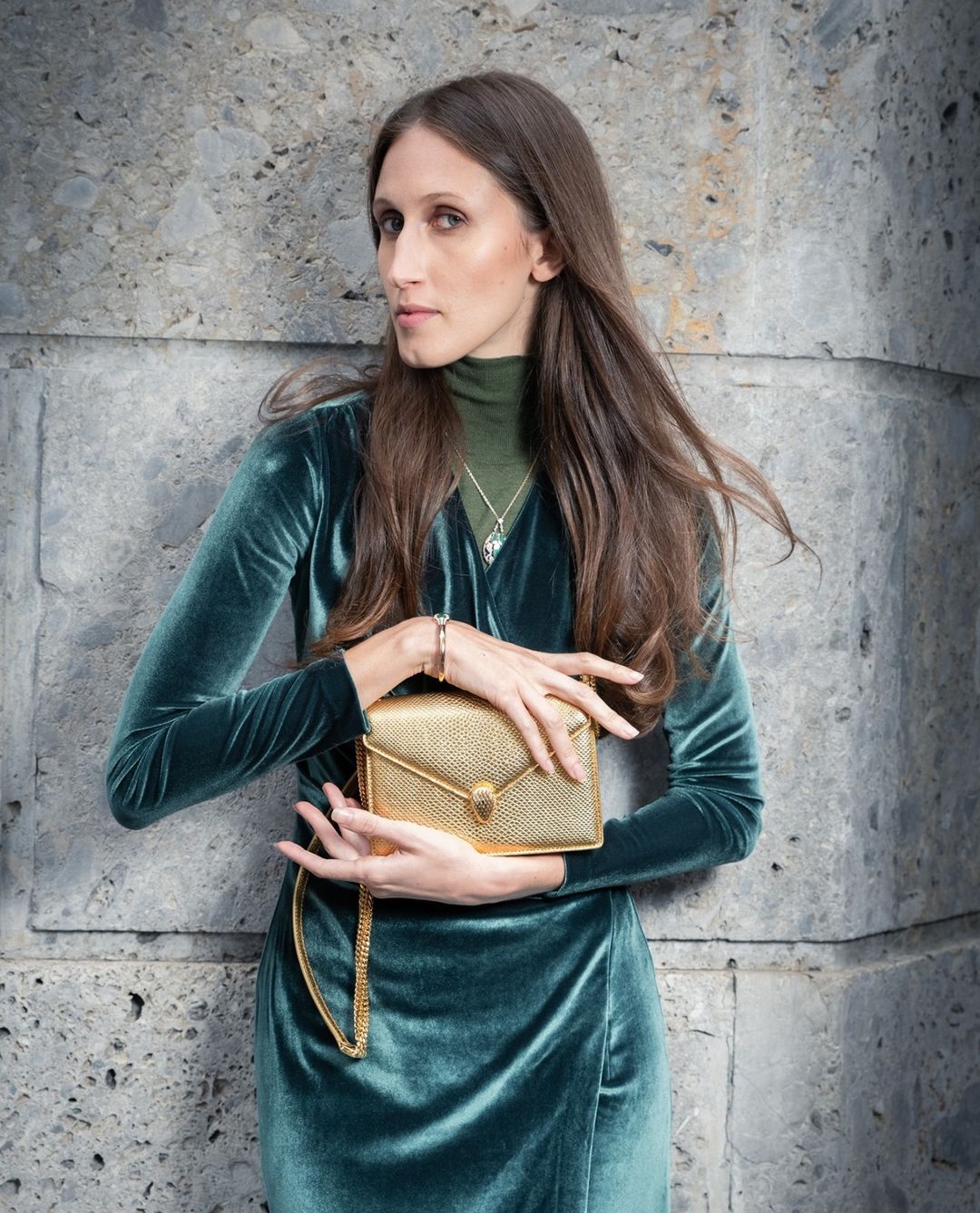 Elegant woman in a green velvet dress holds a gold clutch purse against a textured grey wall, showcasing fashion and style.