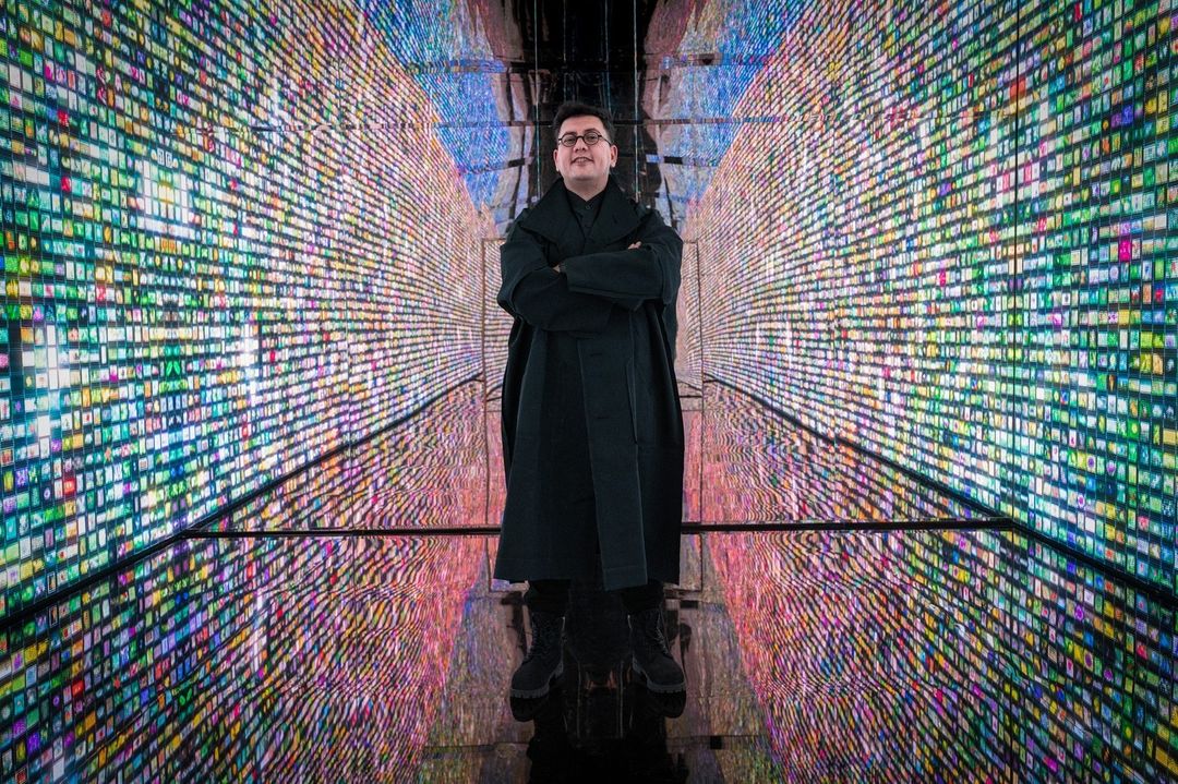 Adult male with arms crossed stands indoors amid colorful digital display walls, creating a futuristic, immersive, and tech-forward scene.