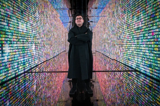 Adult male with arms crossed stands indoors amid colorful digital display walls, creating a futuristic, immersive, and tech-forward scene.