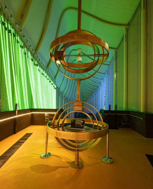 Hourglass encased in a bronze, spherical structure inside of a corridor with green and blue LED lighting and arched design elements.