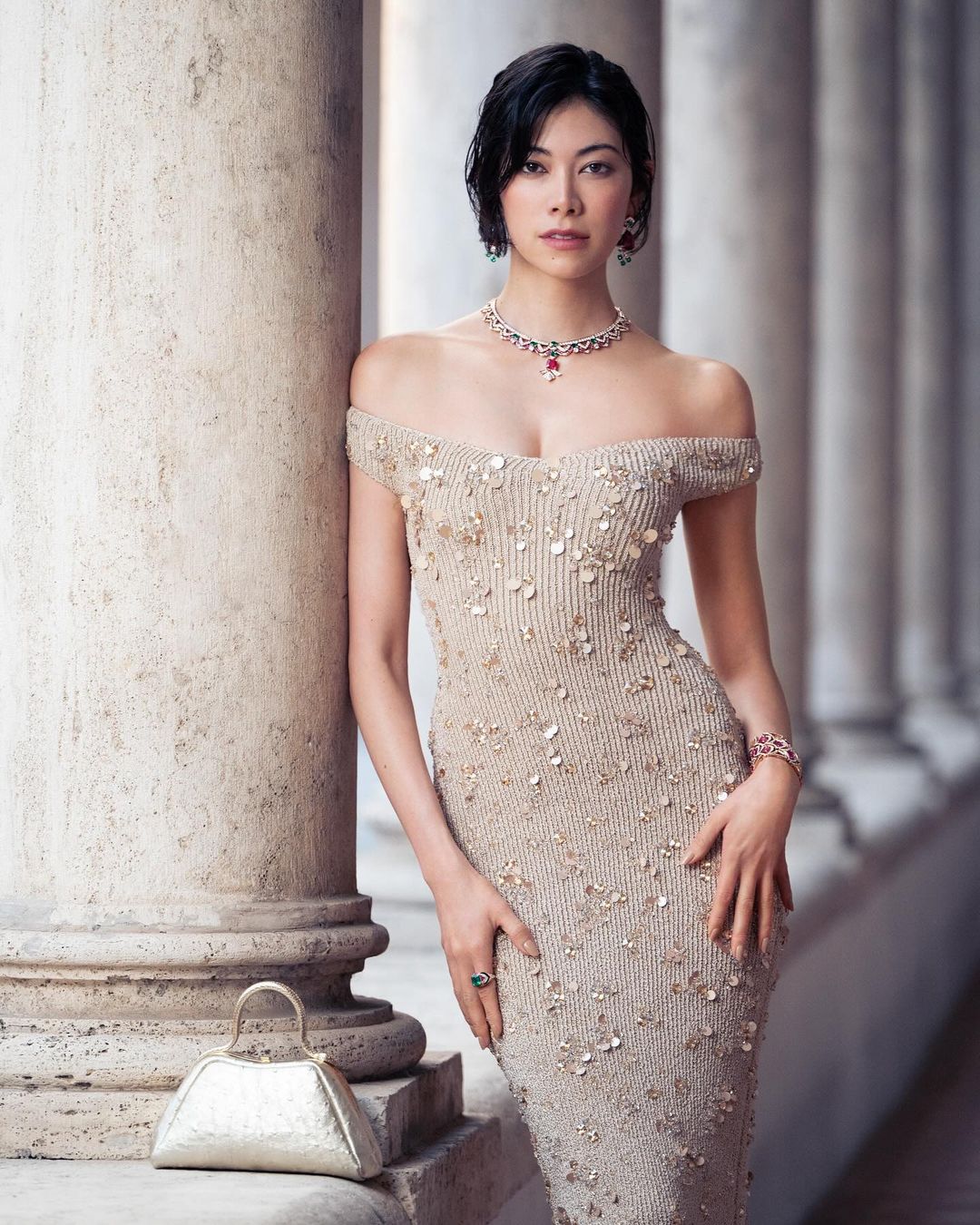 A woman in an elegant beige dress poses gracefully next to a stone pillar, complemented by a metallic purse and jewelry, in a fashion editorial.