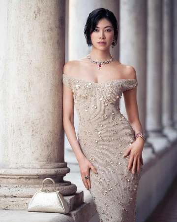 A woman in an elegant beige dress poses gracefully next to a stone pillar, complemented by a metallic purse and jewelry, in a fashion editorial.