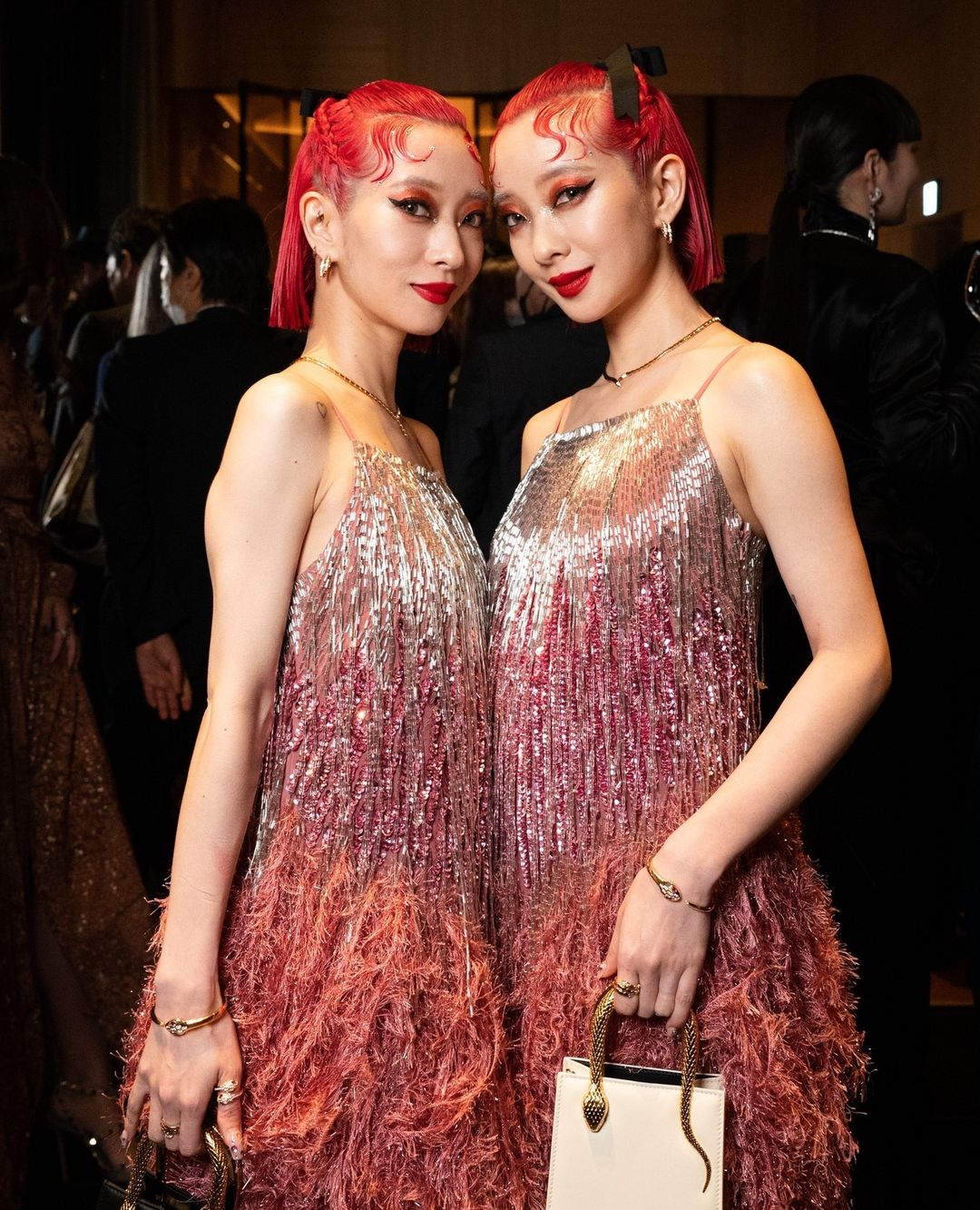 Two adult women with red hair in matching silver and pink fringed dresses hold handbags at an indoor event with a blurred background.