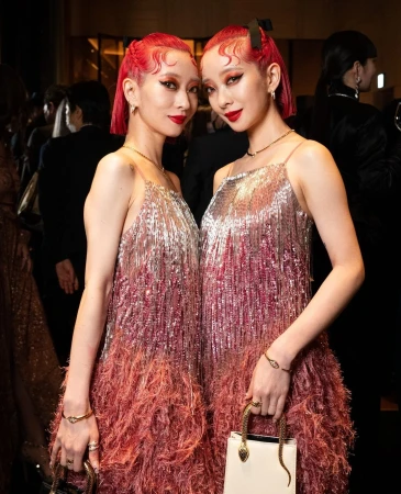 Two adult women with red hair in matching silver and pink fringed dresses hold handbags at an indoor event with a blurred background.
