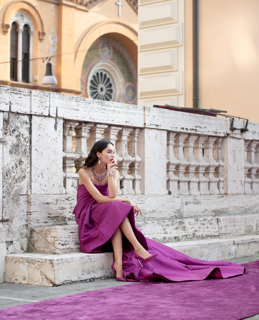 A woman in an elegant purple dress and matching jewelry sits on stone steps in an outdoor setting with a church in the background.