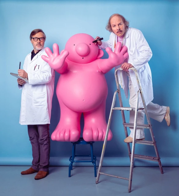 Two men in lab coats pose beside a large pink figure, one on a ladder appearing to draw on the figure in a studio setting.