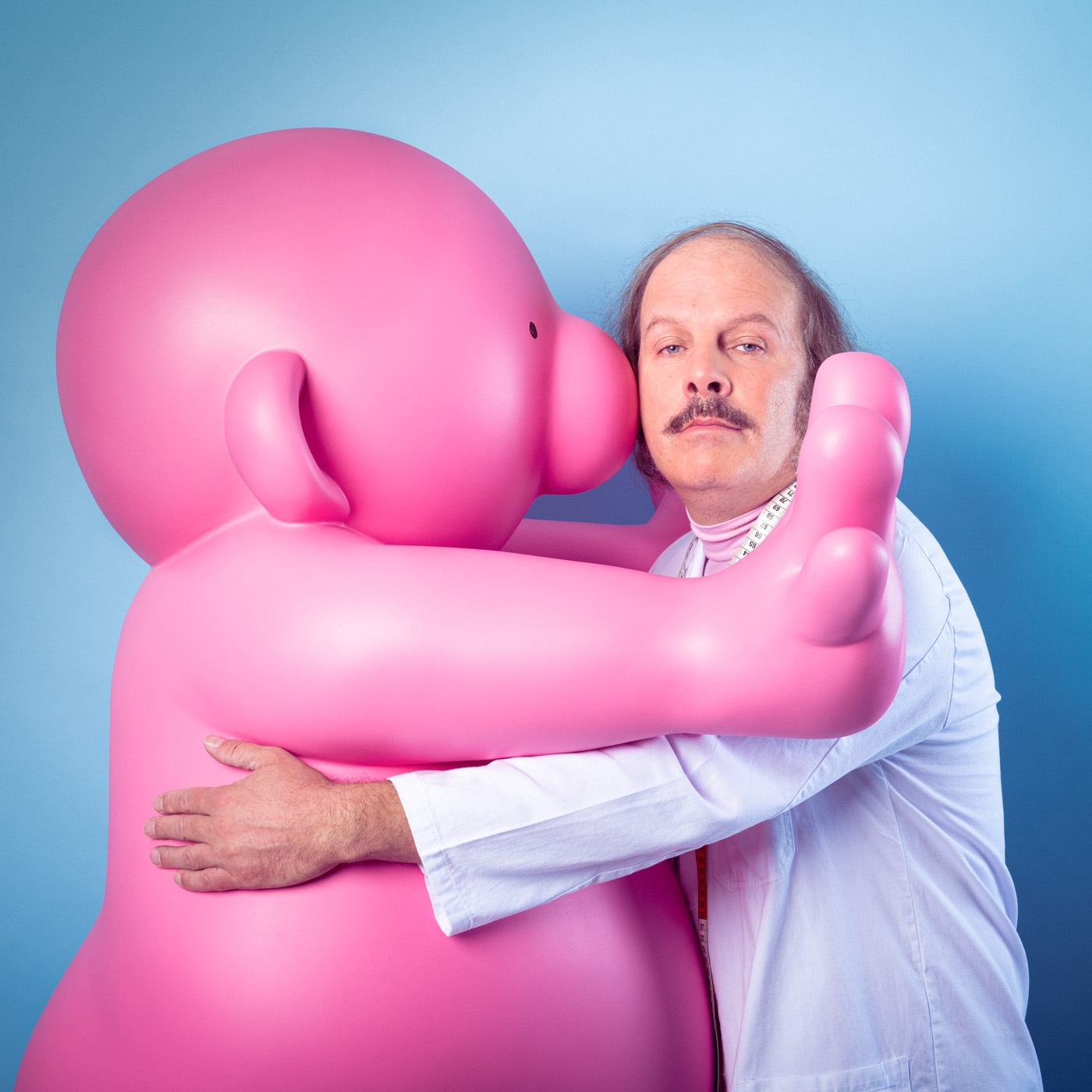 Man in white coat embraces a large pink cartoon figure against a blue background, creating a whimsical and eye-catching portrait.